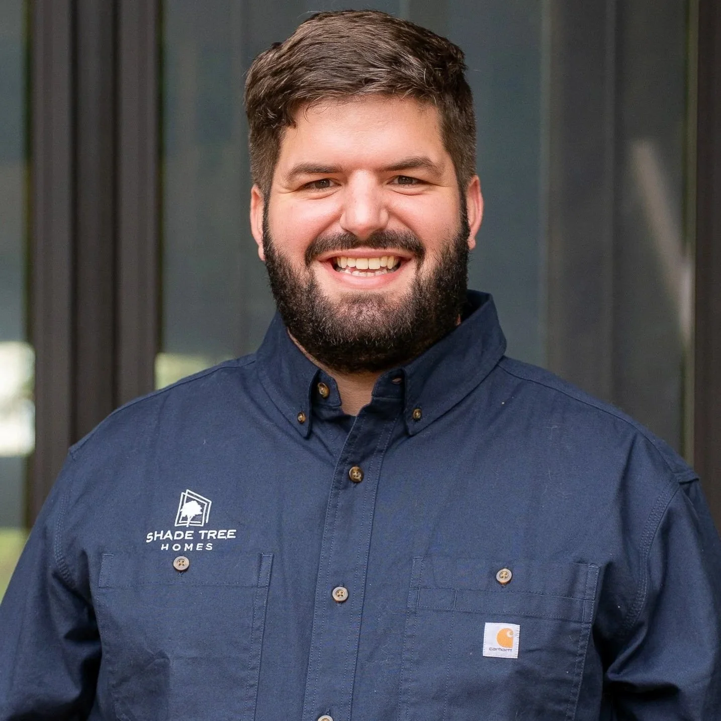 Portrait of a happy man with short dark hair and a beard, wearing a navy blue work shirt with a company logo, standing outdoors in front of a building with glass windows.
