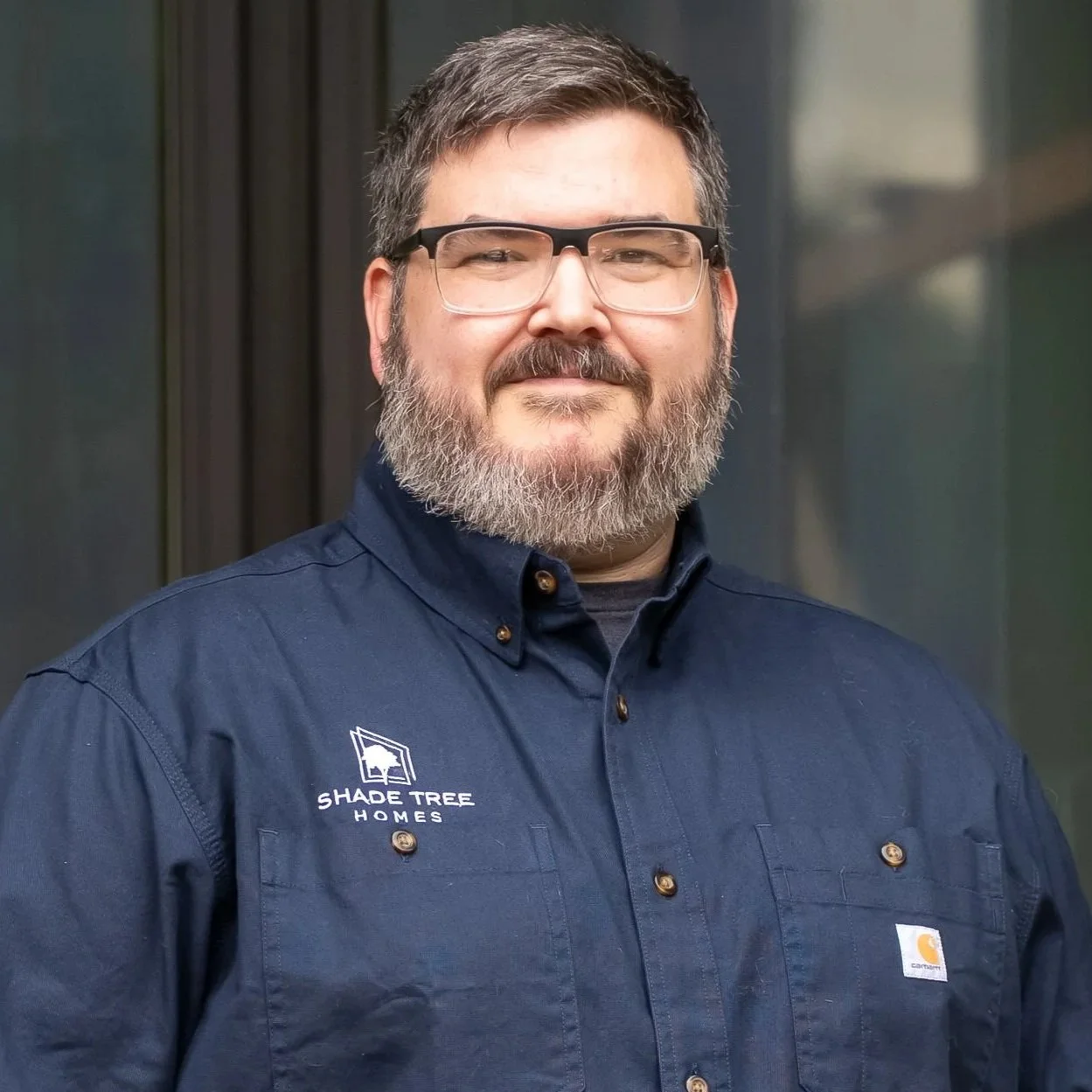 A man with glasses, a beard, and gray hair smiling, wearing a navy blue shirt with the logo 'Shade Tree Homes' and a Carhartt logo, standing in front of a dark, blurred background.