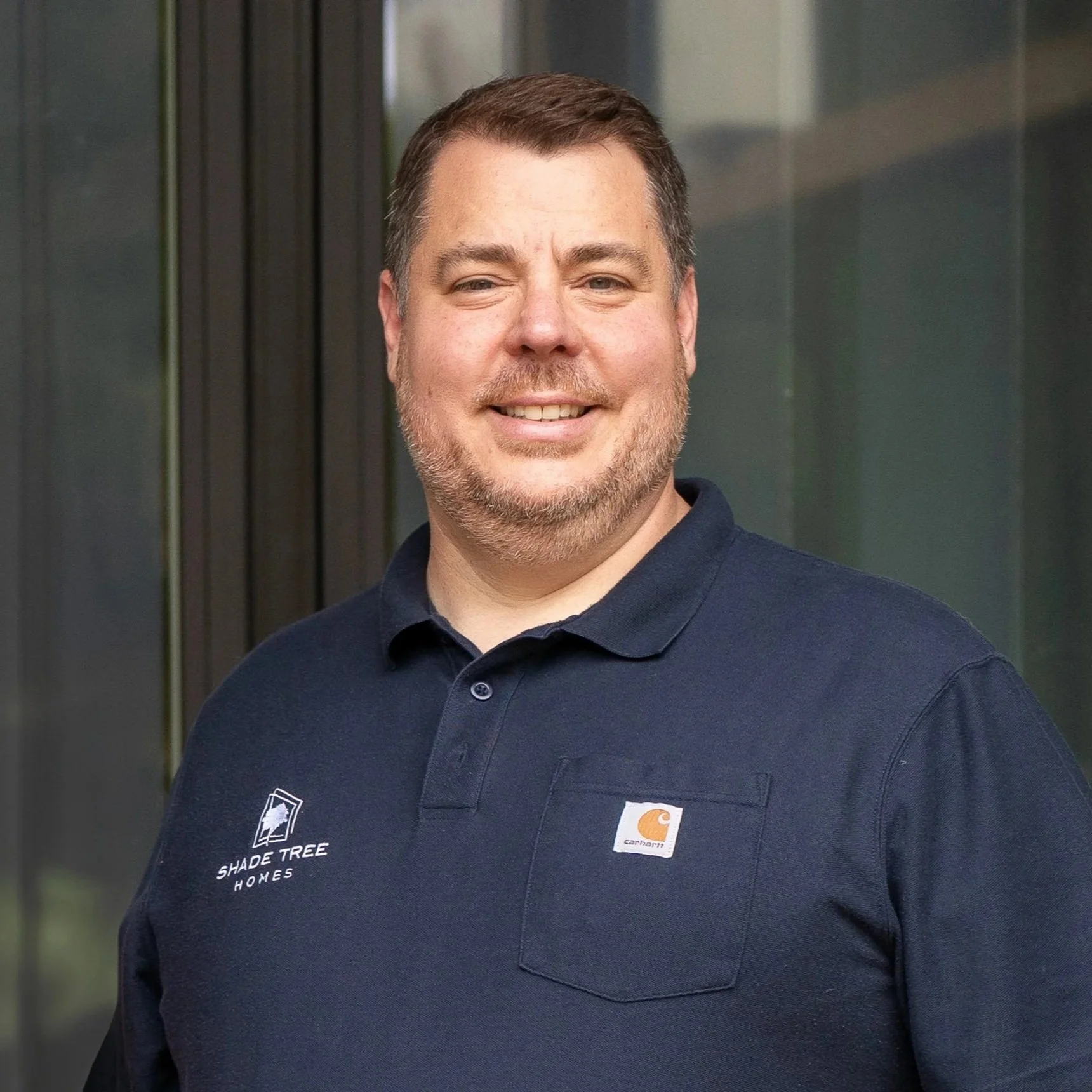 A man smiling outdoors, wearing a dark blue Carhartt polo shirt with a small logo patch on the right side and a Shade Tree Homes logo on the left side.