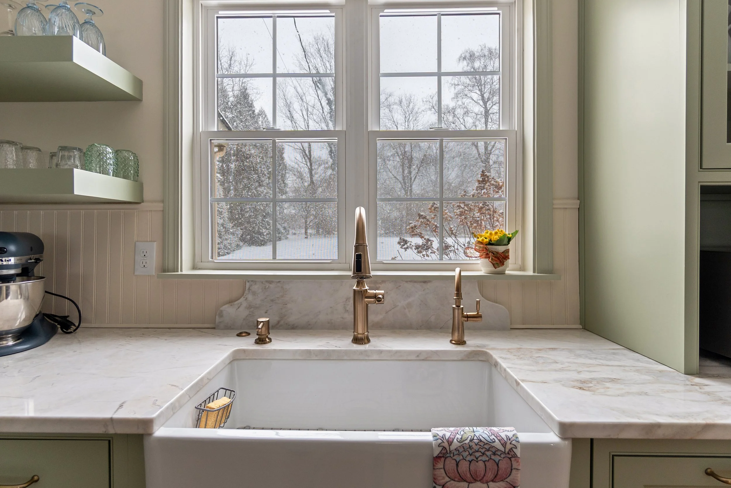 Kitchen with white marble countertop, modern copper faucet, window overlooking snowy landscape, green cabinets, open shelving with glassware, a plant, and a towel hanging on the sink.