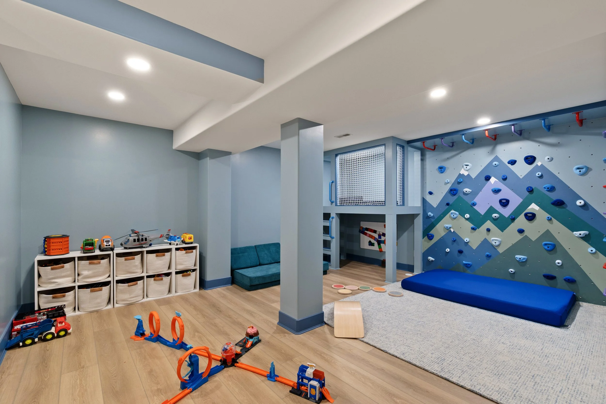 Children's play area with toy vehicles, a climbing wall, and a cozy seating area in a room with light blue walls and wooden flooring.