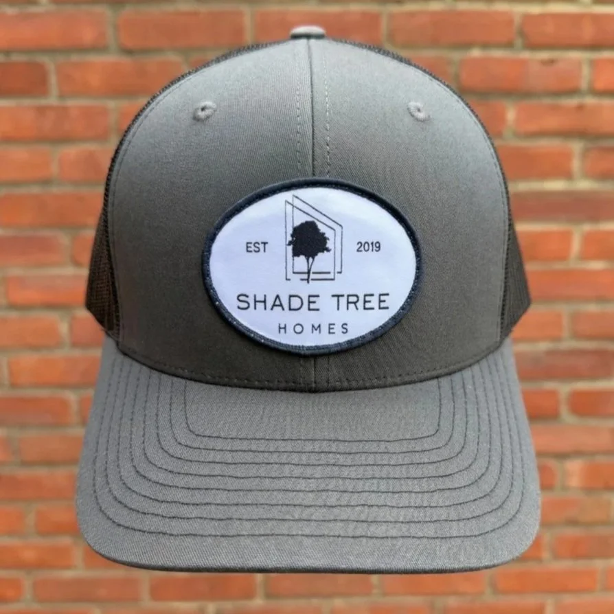 Gray and black trucker hat with a patch featuring a tree, a door outline, and the text 'EST 2019 SHADE TREE HOMES' against a brick wall background.