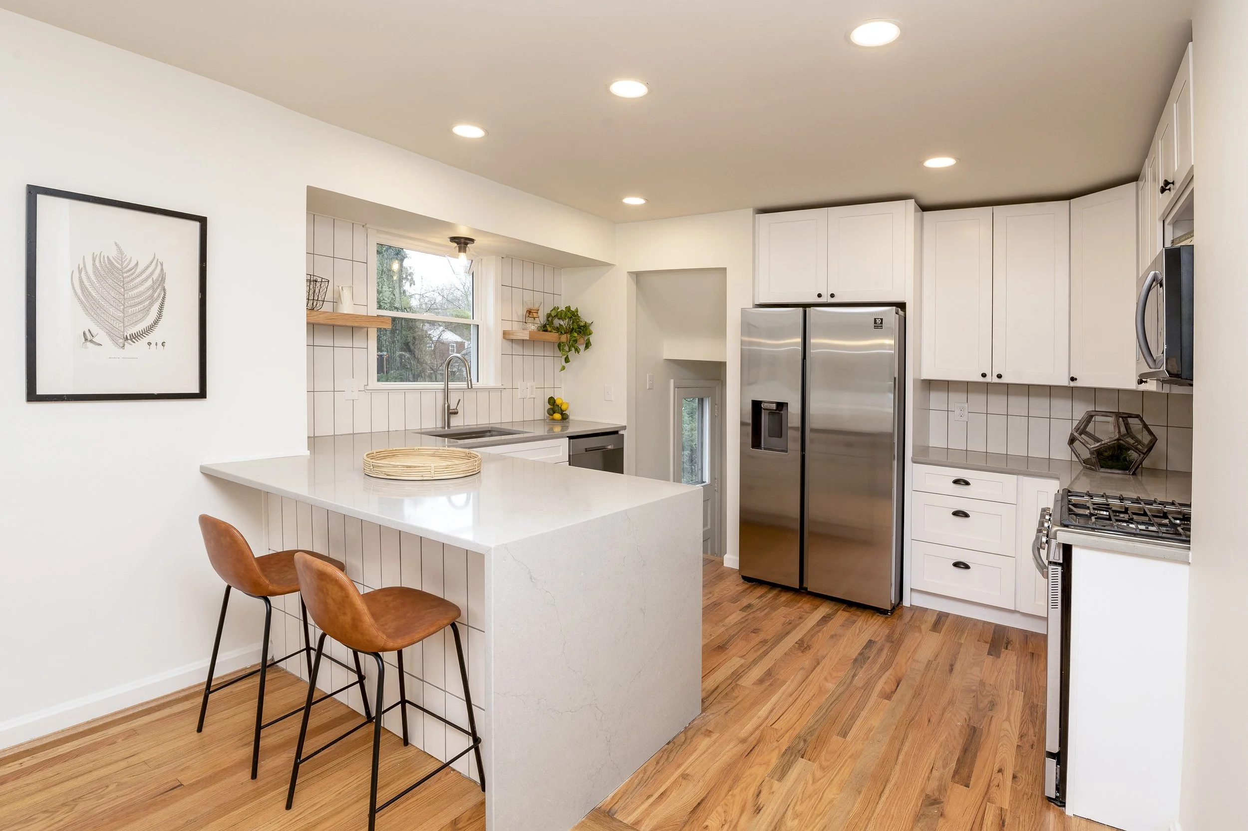 A modern kitchen with white cabinets, stainless steel refrigerator, and a white countertop island with two brown leather barstools. The kitchen has hardwood floors, a window with a view outside, and minimal decor.