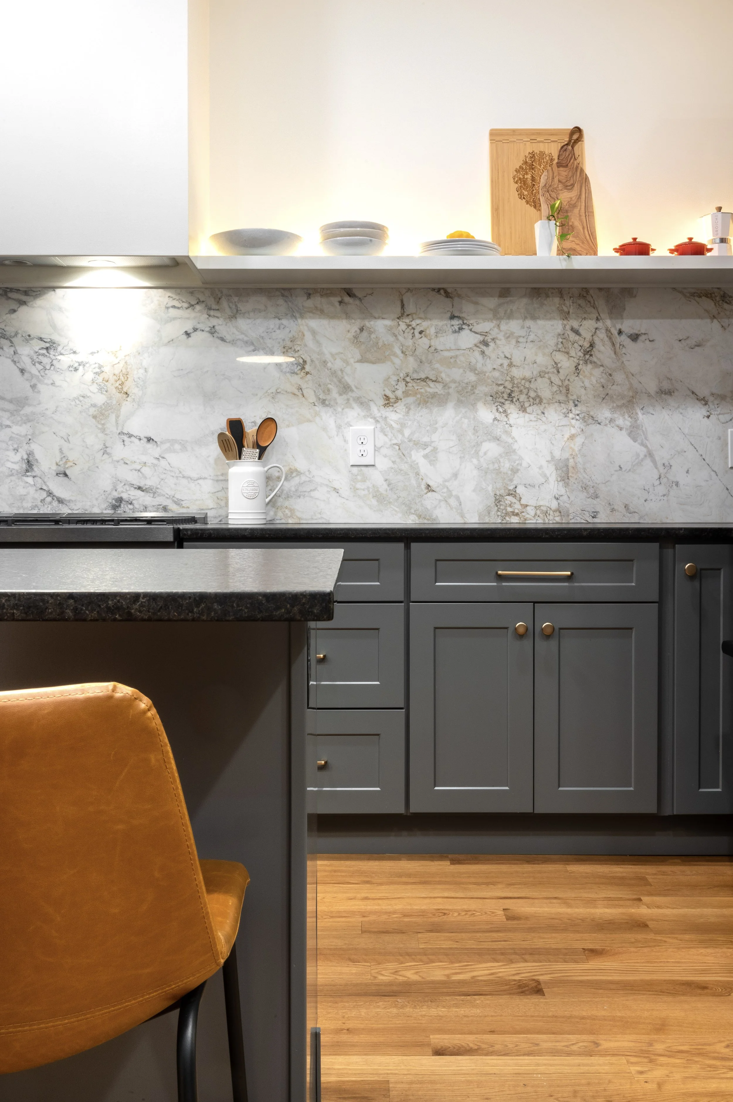 View of a kitchen countertop and cabinets with a marble backsplash. The countertop has a utensil holder with wooden and black utensils, and a power outlet is visible on the wall. An open shelf above displays bowls, a cutting board, a small plant, and red pots. A tan chair is partially visible in the foreground.