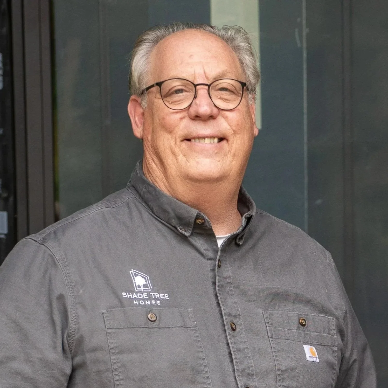A middle-aged man with gray hair and glasses smiling at the camera, standing in front of a dark background. He is wearing a dark gray shirt with a 'Shade Tree Homes' logo on the left chest.