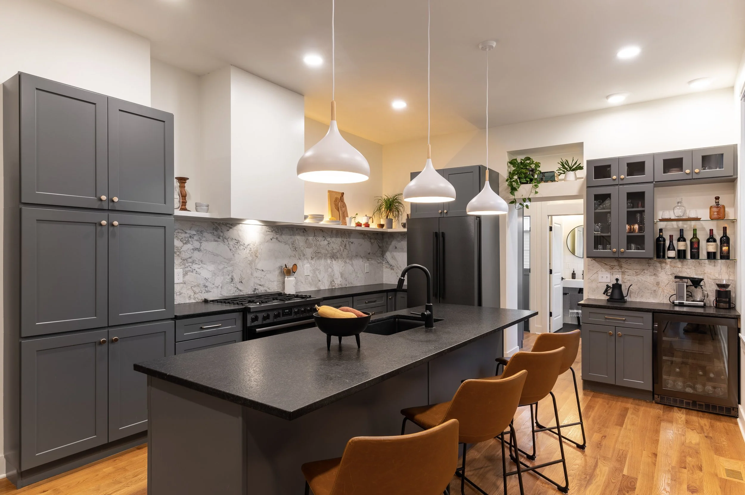 Modern kitchen with grey cabinets, black countertops, a marble backsplash, and pendant lights over the island; there are plants and bottles on display.