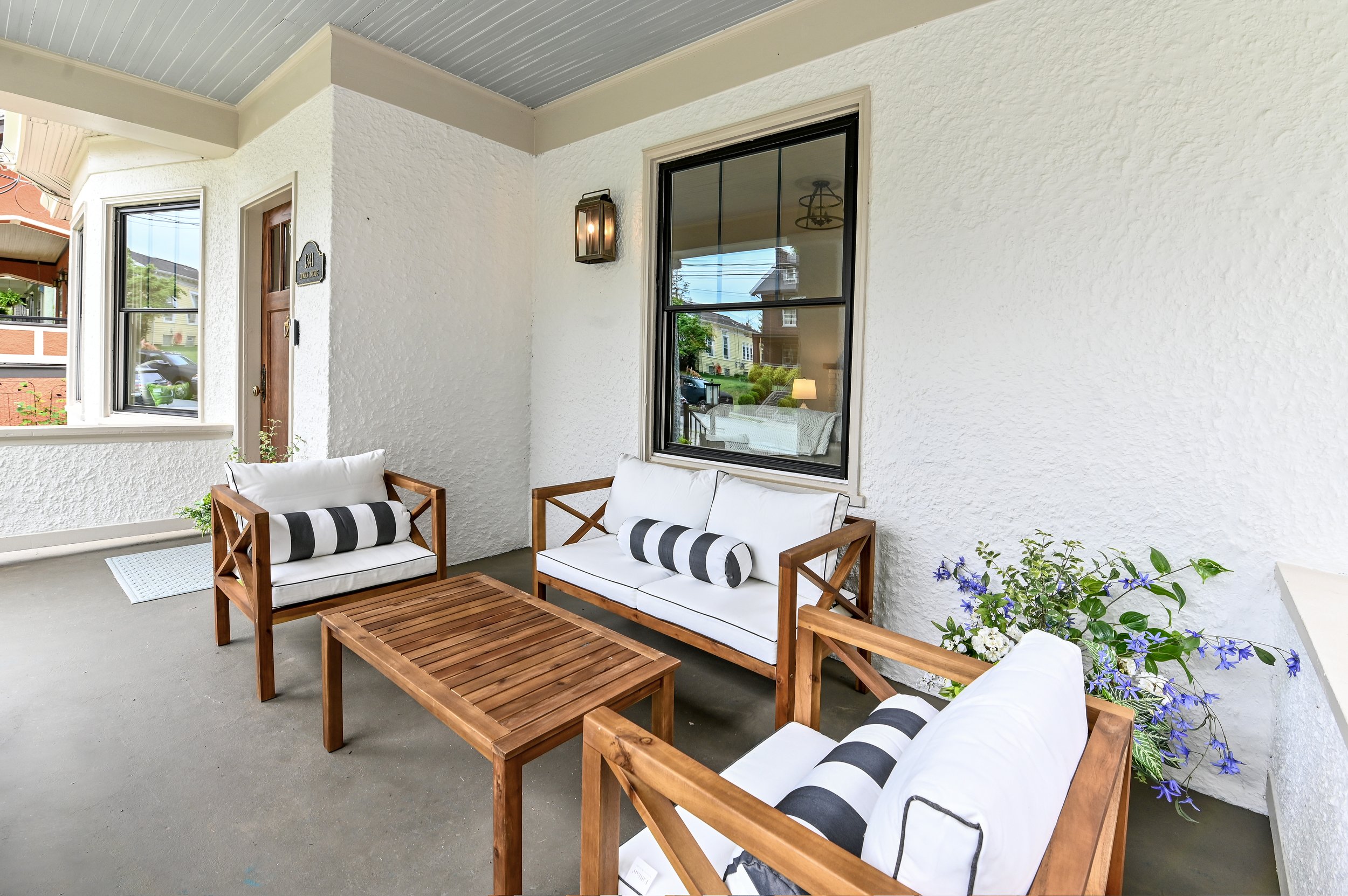Exterior porch with white textured wall, black framed windows, wooden outdoor furniture with white cushions and striped pillows, potted purple flowers, and a wall-mounted lantern light.