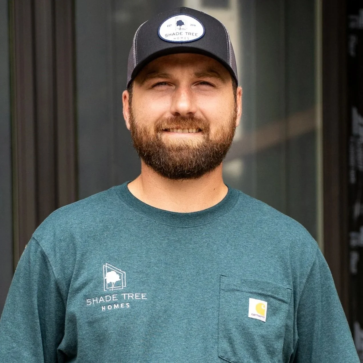 A smiling man with a beard wearing a black and gray Shade Tree Homes baseball cap and a teal Carhartt t-shirt. He is standing outside against a dark-colored building.