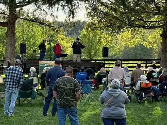 Group of people gathered outdoors listening to three speakers on a small stage surrounded by trees