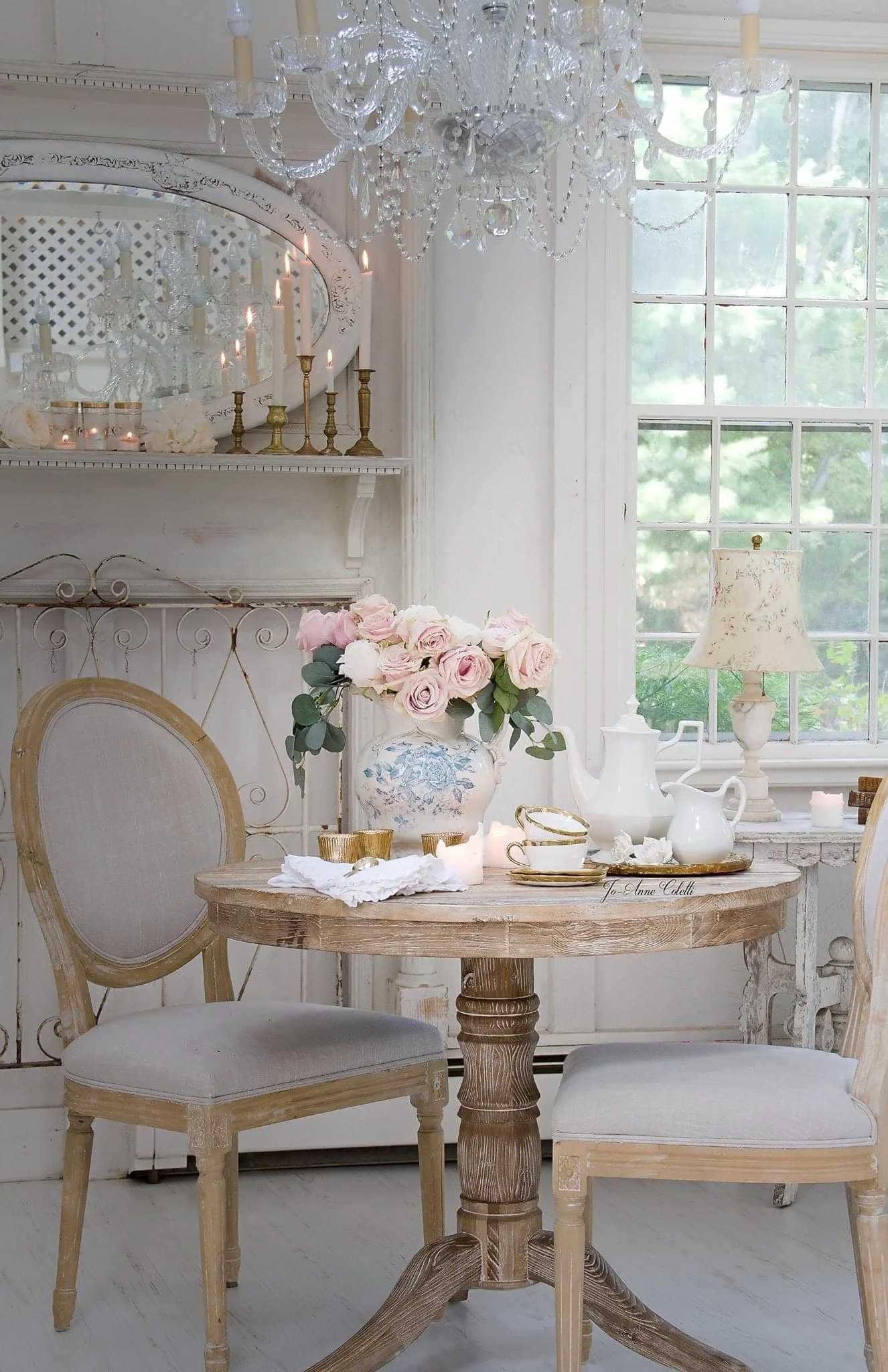 A cozy, elegant dining nook in a bright room with large window, vintage wooden table, upholstered chairs, floral bouquet in a vase, white dishes, gold-rimmed cups, teapot, and a white lamp on a side table. Crystal chandelier hanging from ceiling.