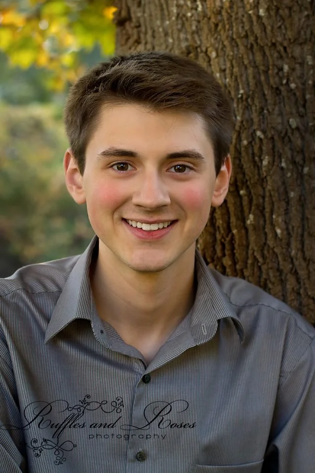A young man with brown hair and wearing a gray button-up shirt, smiling in front of a tree with green foliage in the background.