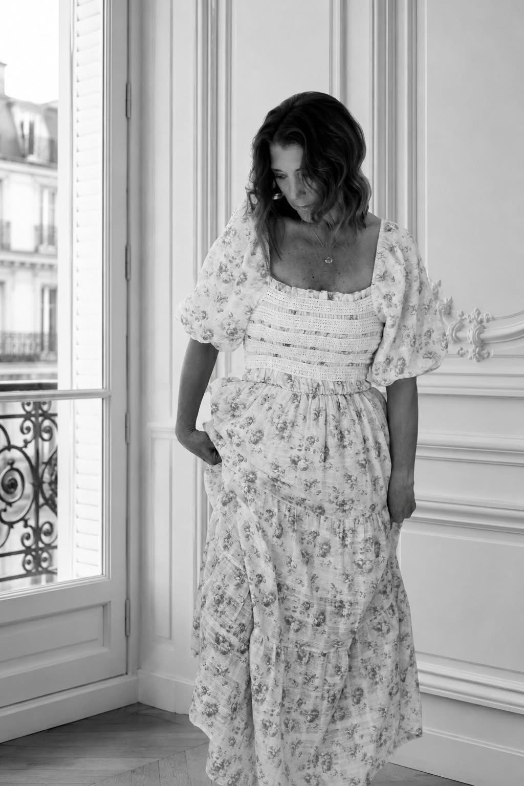 A woman in a floral dress standing near a window with a view of a building, in a room with ornate wall molding.