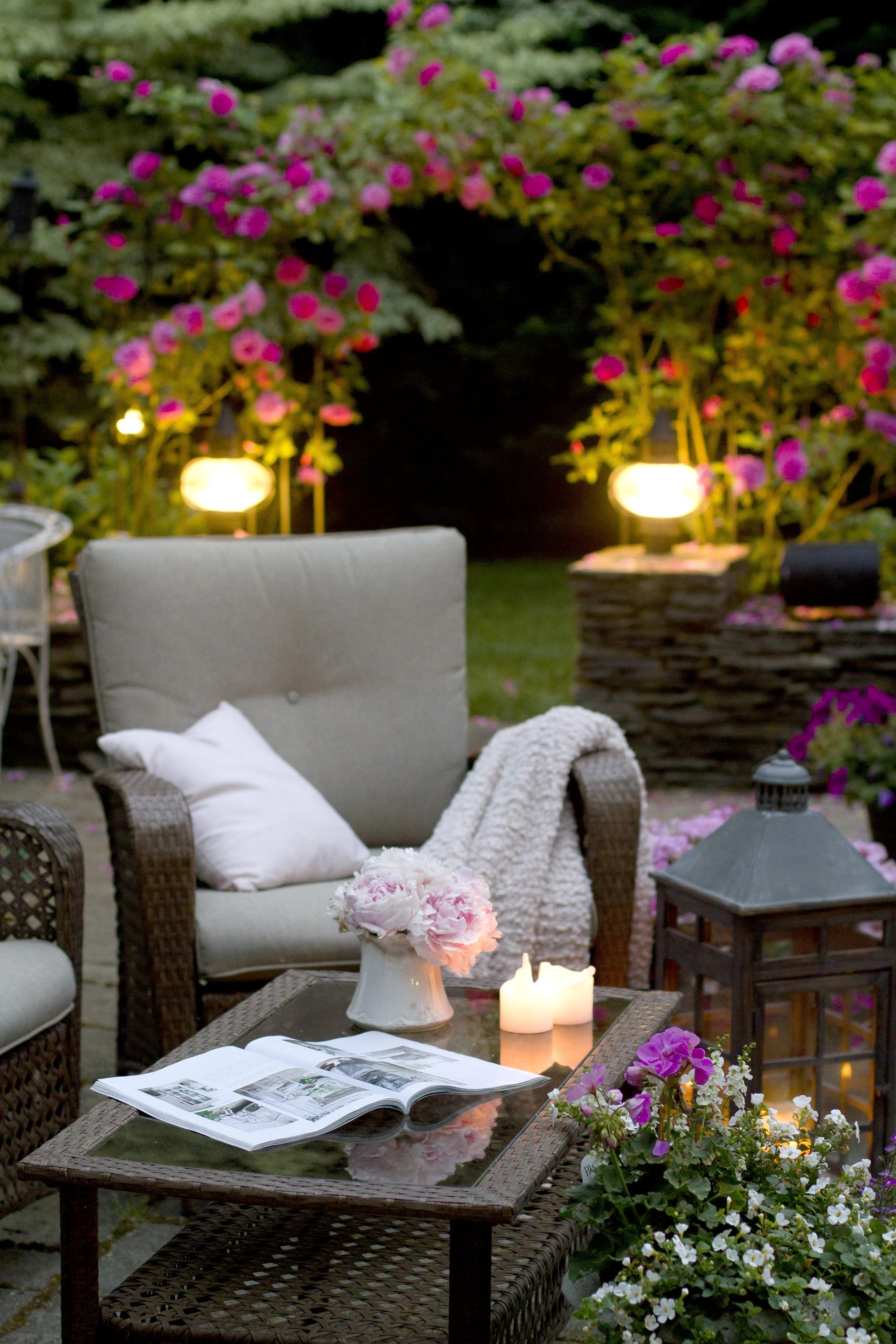 An outdoor patio evening scene with wicker furniture, a glass-topped table, candles, a vase of pink peonies, and blooming pink and white flowers, with lanterns in the background.