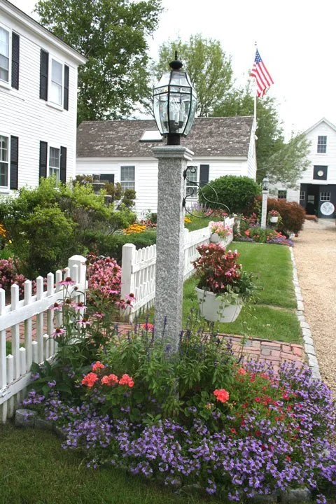 A garden scene with a white picket fence, colorful flowers, a stone lamp post, a white house with black shutters, and an American flag on a pole.