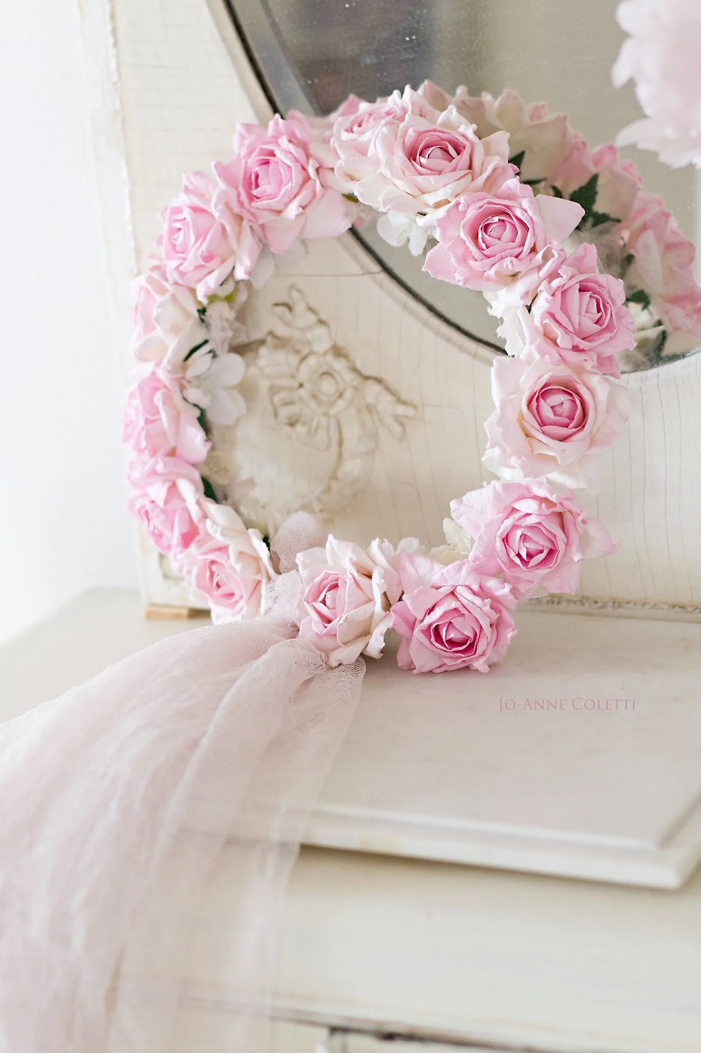 A floral wreath made of pink roses and greenery, resting on a white surface near a mirror.