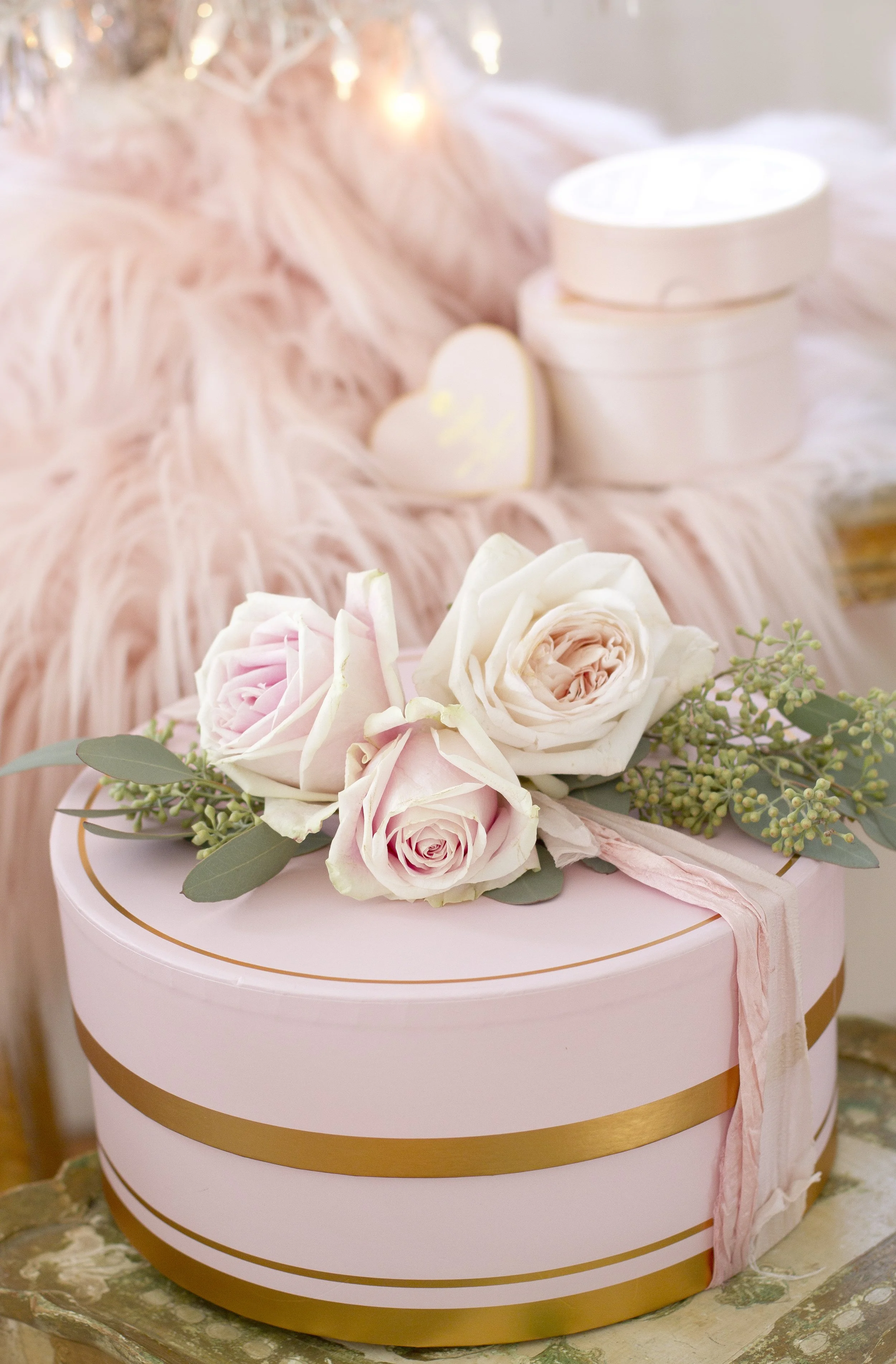 A pink round gift box decorated with a light pink satin ribbon, topped with a bouquet of pale pink and white roses and greenery, placed on a vintage-style tray.