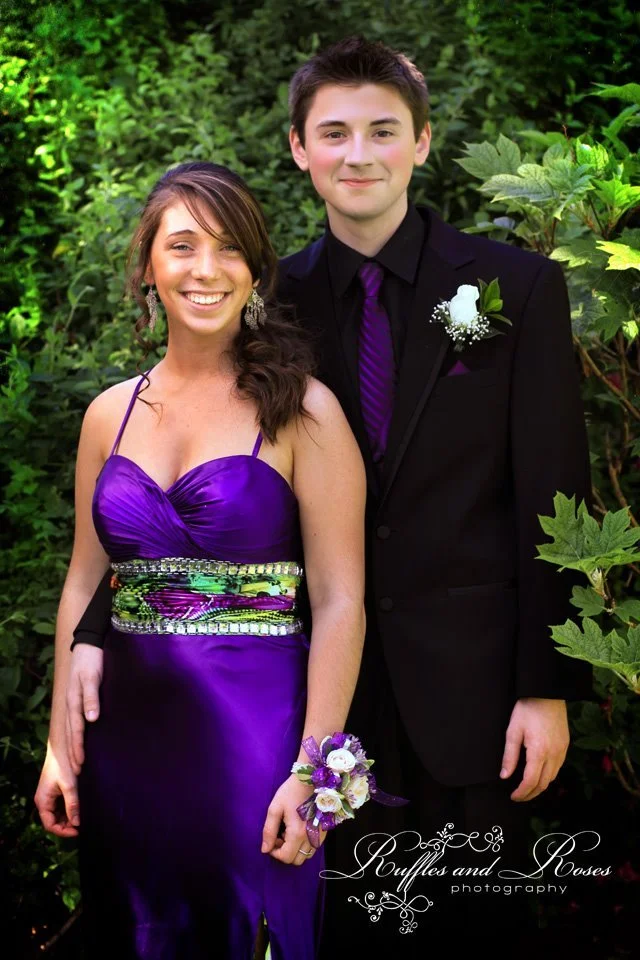 A young woman in a purple dress and a young man in a black suit with a purple tie stand together outdoors near green foliage, smiling at the camera.