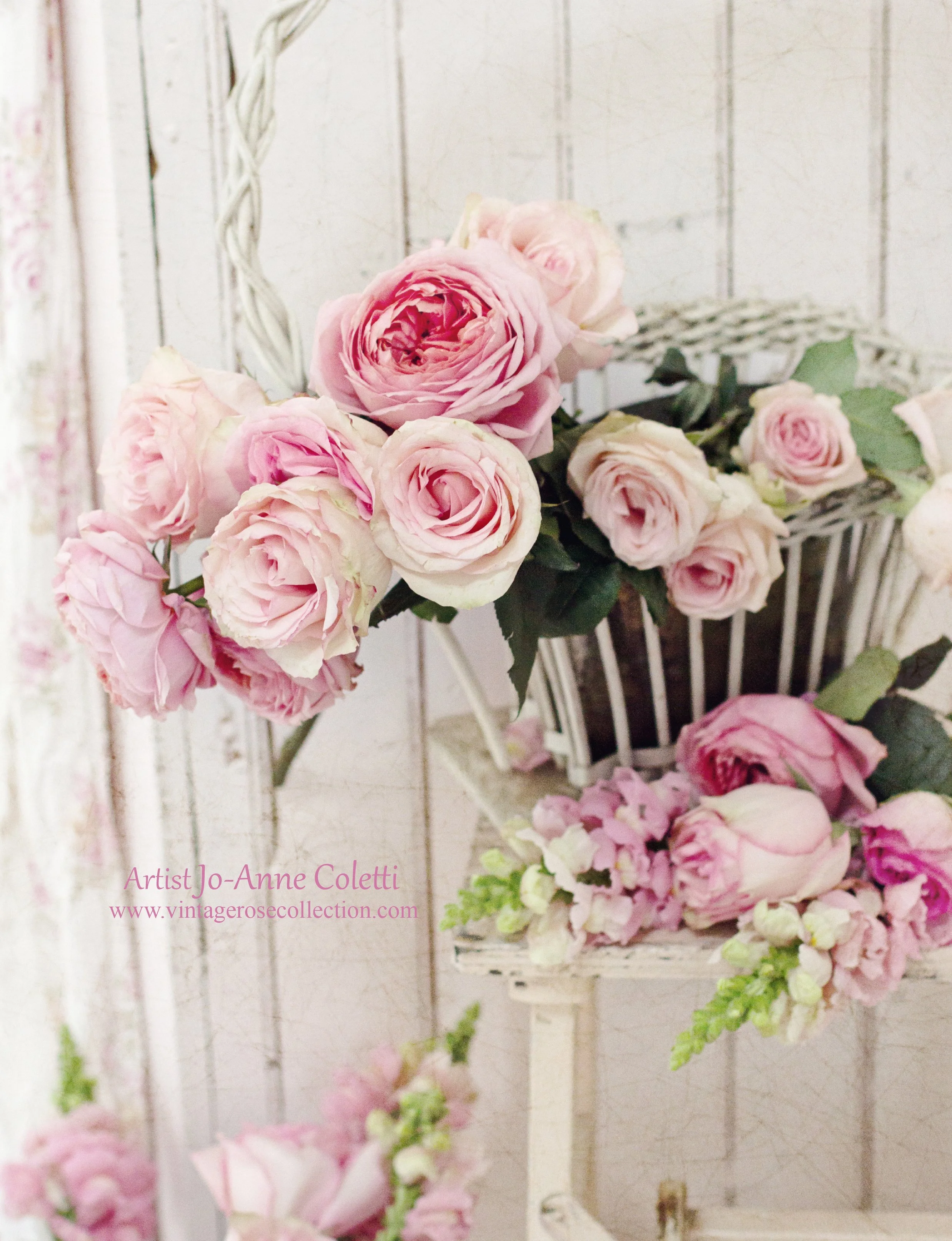 Pink and cream roses arranged in a white wicker basket with a white wooden background and some greenery.