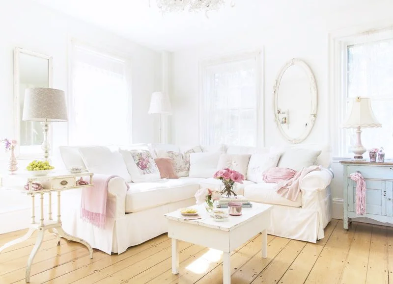 Bright, white living room with a large L-shaped sofa adorned with pink and floral cushions, a white coffee table with a pink flower arrangement, and vintage-style lamps on side tables, set on light wood flooring and decorated with a large oval mirror