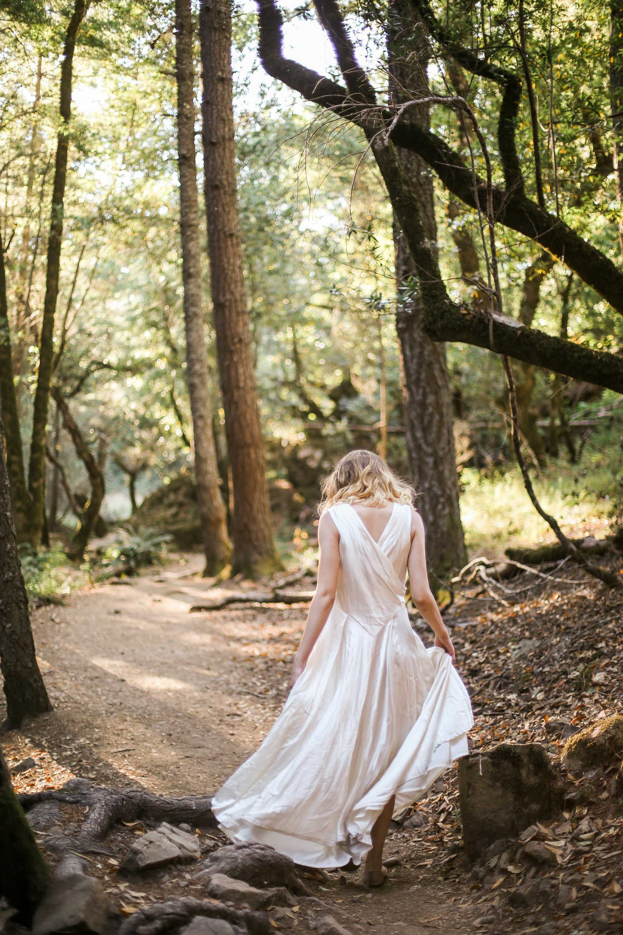 A woman in a white dress walking through a wooded forest path with trees and sunlight filtering through the leaves.