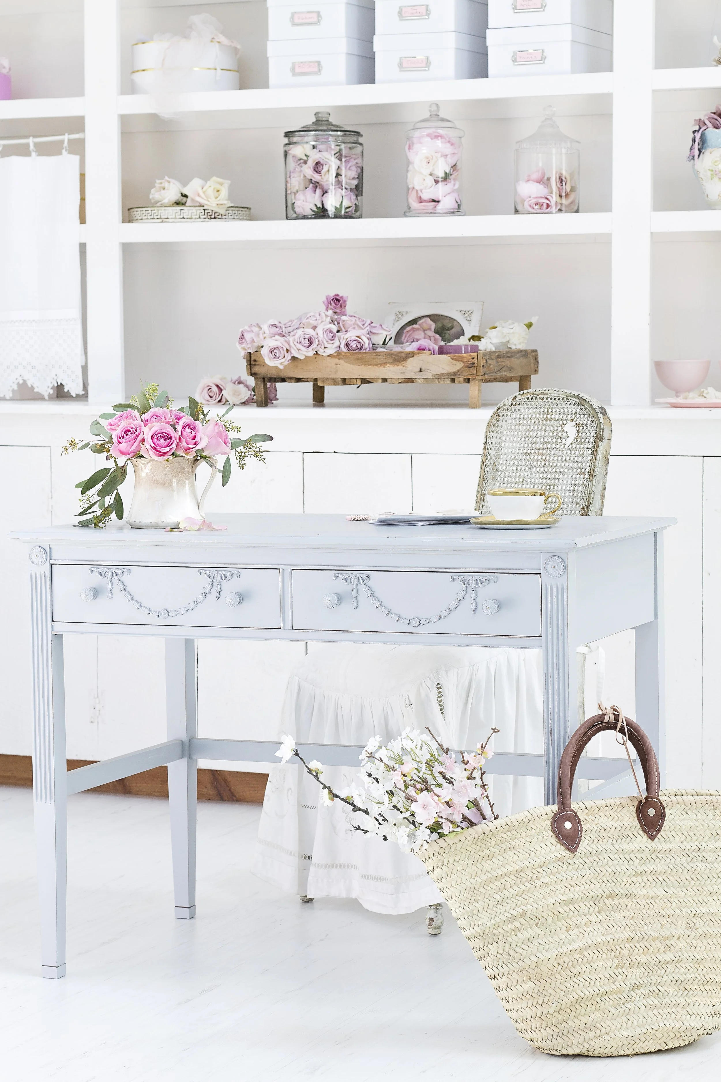 A white vintage-style dresser with floral decorations, a branch of pink blossoms, and a woven straw bag on a white floor in a bright room.
