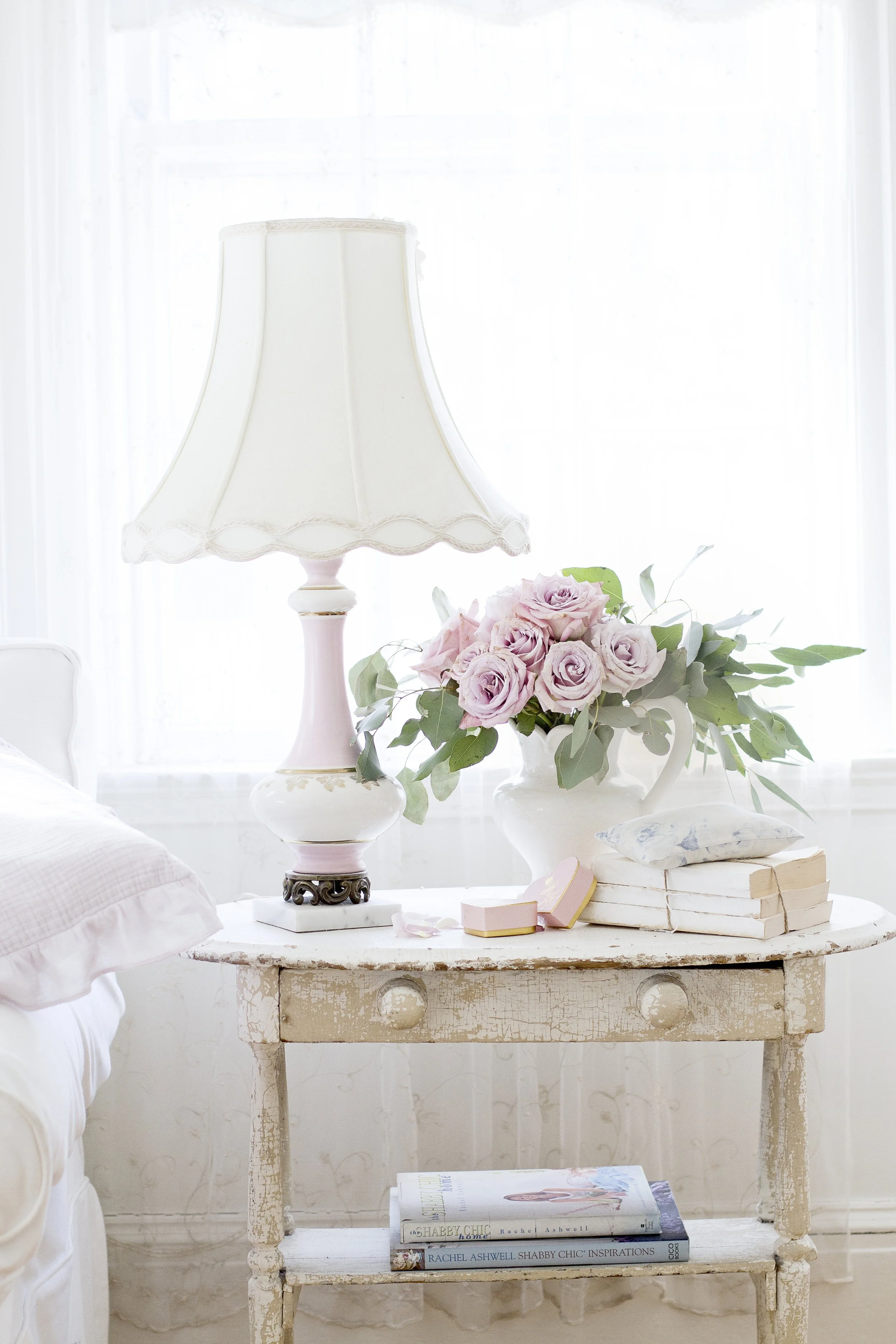 A vintage white wooden table with a distressed finish holding a stack of books, a floral arrangement of pink roses and green leaves in a white pitcher, a pink jewelry box, and a decorative peach-colored heart. On top is a pastel pink and white table 