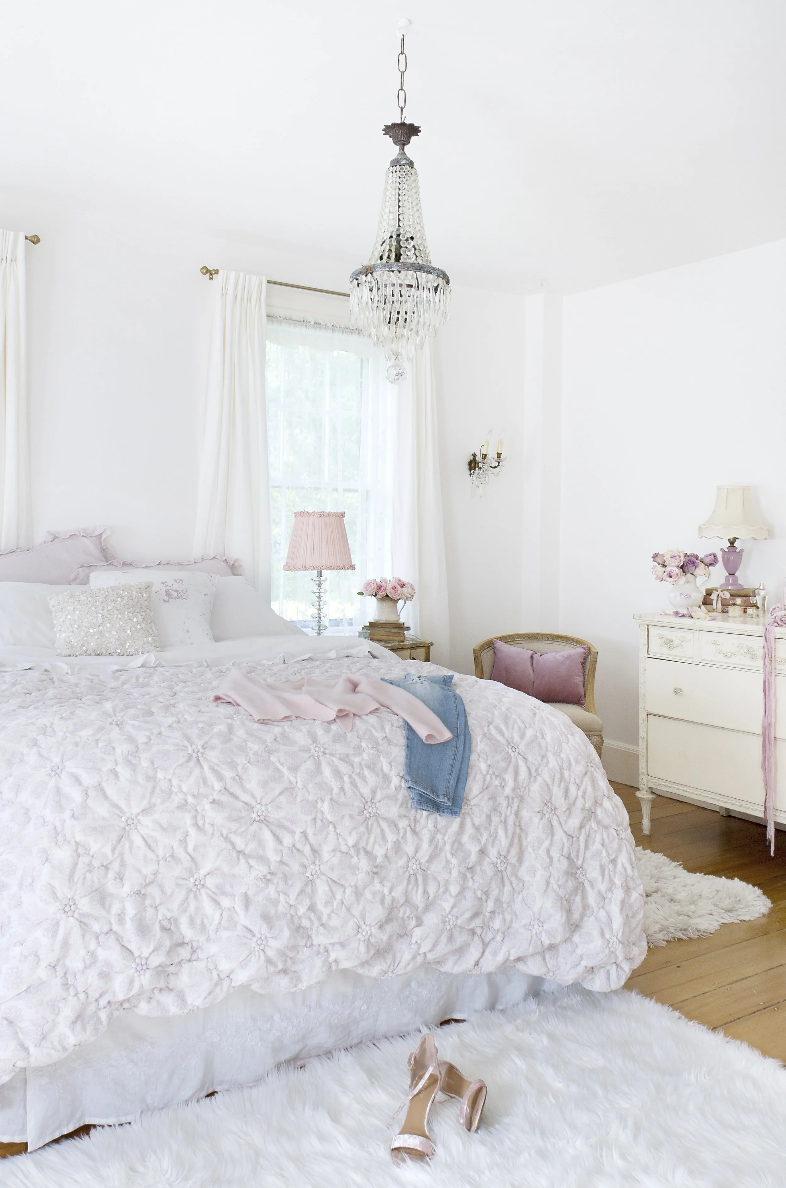 A bedroom with white walls, a white bed with pink and white bedding, a bedside table with pink flowers, a pink lampshade, a pair of nude high heels on a white furry rug, a vintage dresser with floral decor, and a crystal chandelier.