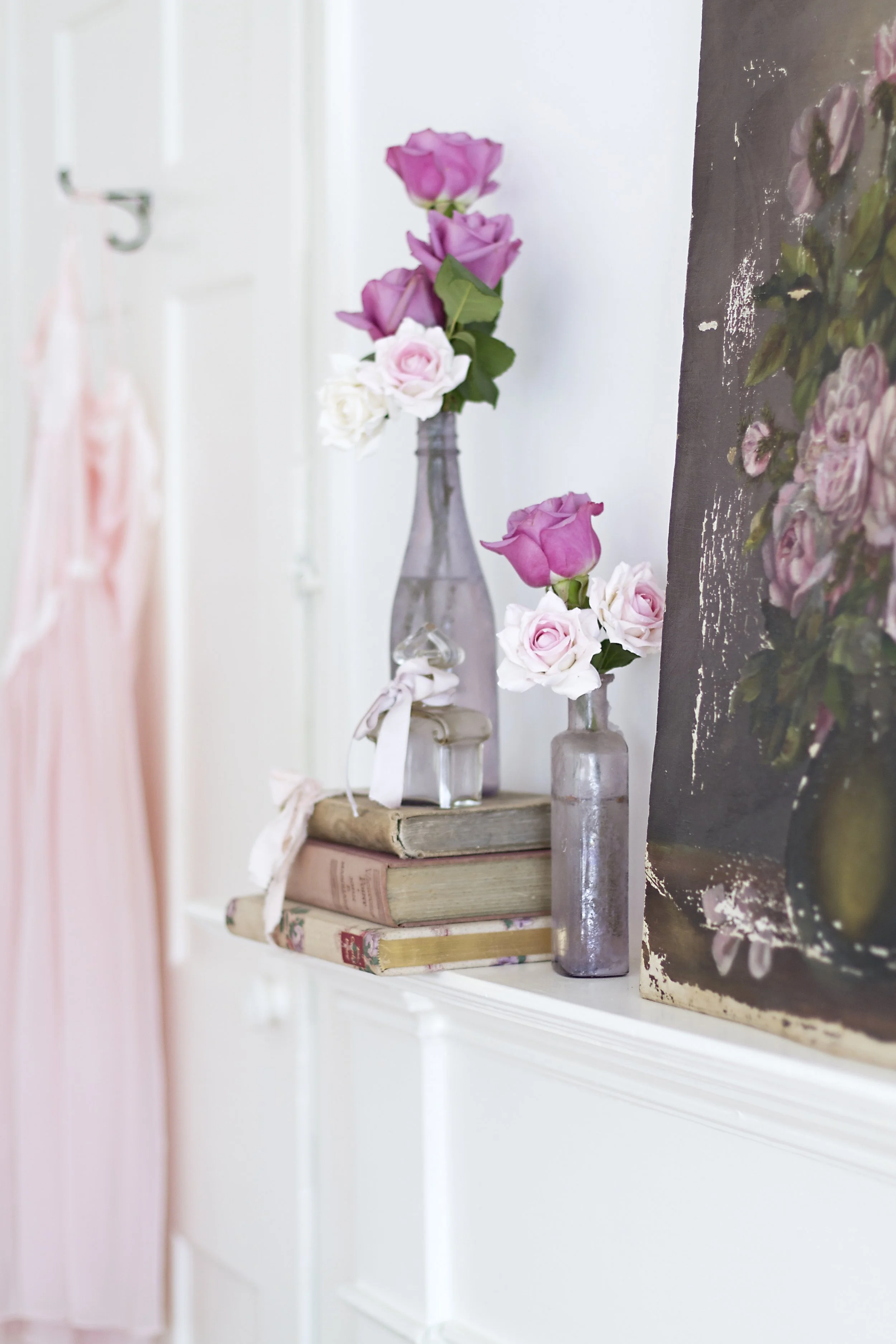 A shelf with vintage books, a glass bottle with pink and white roses, and a painting with floral imagery.