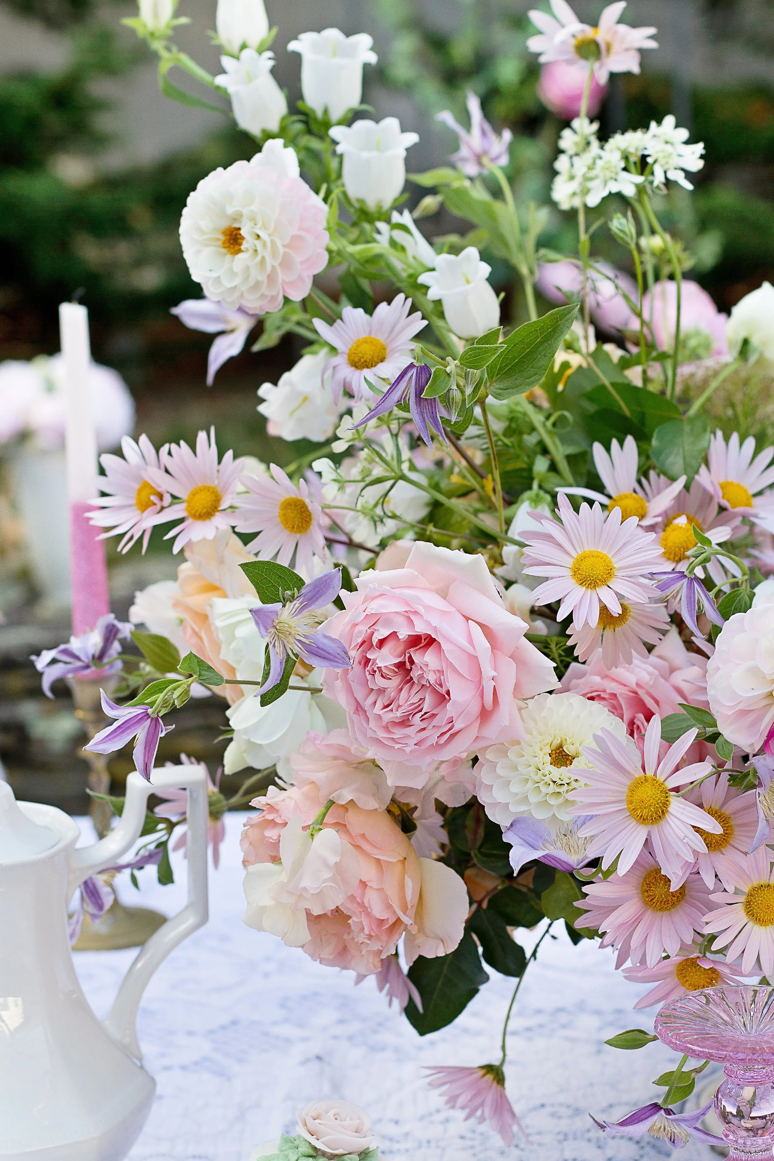 A large, elegant floral bouquet with pink and white roses, daisies, and other flowers arranged on a white table, with a white teapot nearby and a blurred outdoor background.