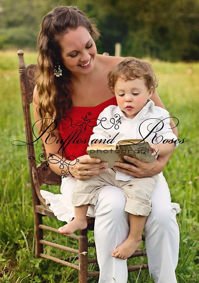 A woman and a young boy sitting outdoors on a wooden chair in a grassy field, looking at a book together.