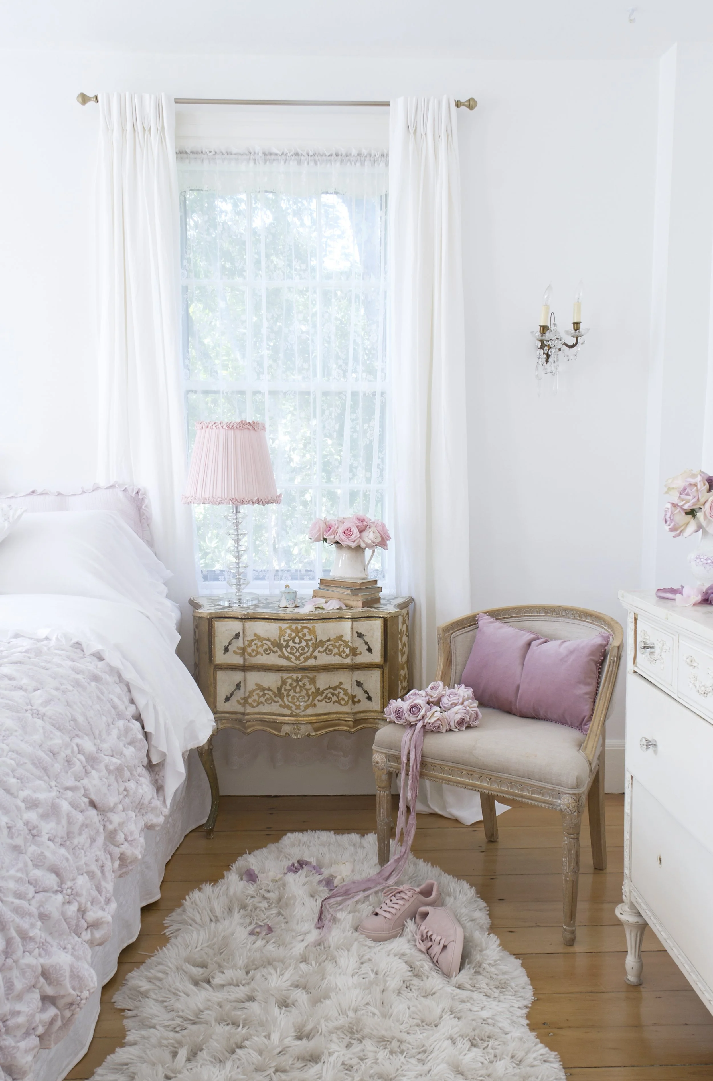 A cozy bedroom with white walls, sheer curtains, a window with greenery outside, a vintage nightstand with a pink lamp and roses, a plush white rug, a beige cushioned chair with pink and purple pillows, a white dresser, and pink sneakers on the rug.