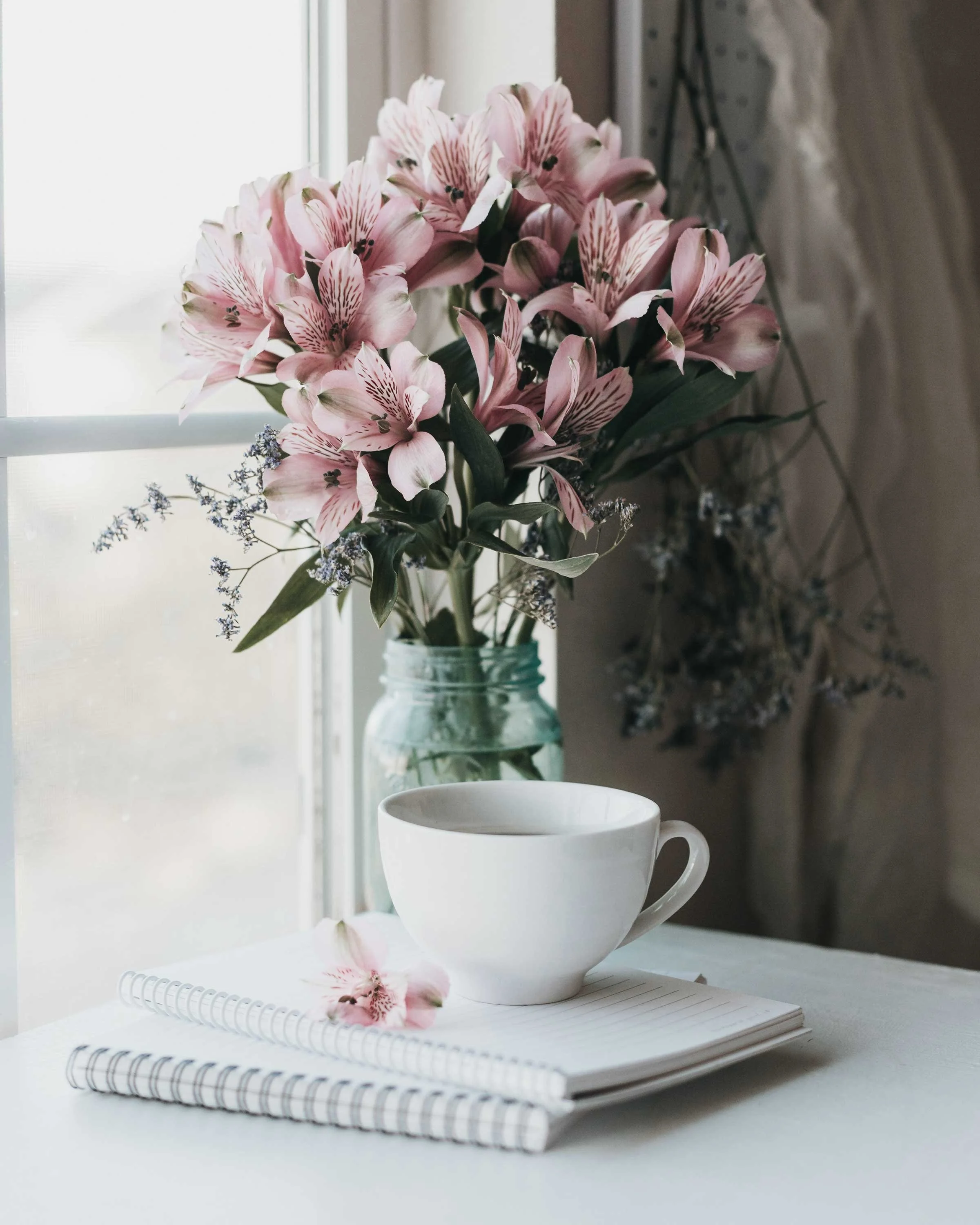 A bouquet of pink lilies in a glass jar on a windowsill, with a white mug and a notebook with a pen on top in the foreground.