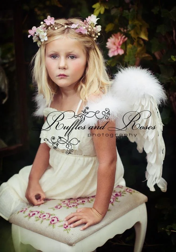 A young girl dressed as an angel with white feathered wings, wearing a white dress and a flower crown, sitting on a cushioned bench outdoors.