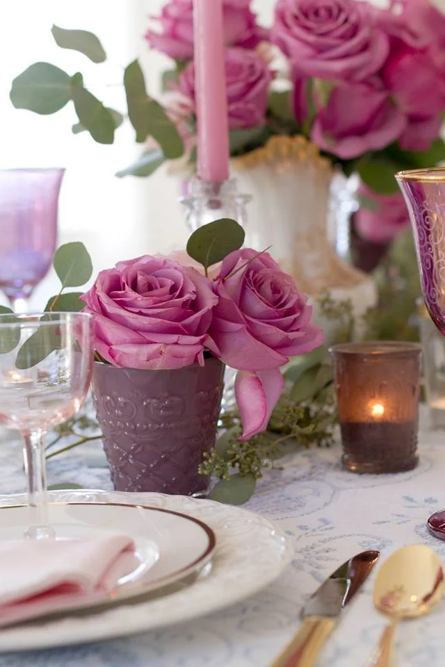 Pink roses in a purple vase on a table decorated with pink, purple, and gold candles, a white plate with a pink napkin, and gold cutlery.