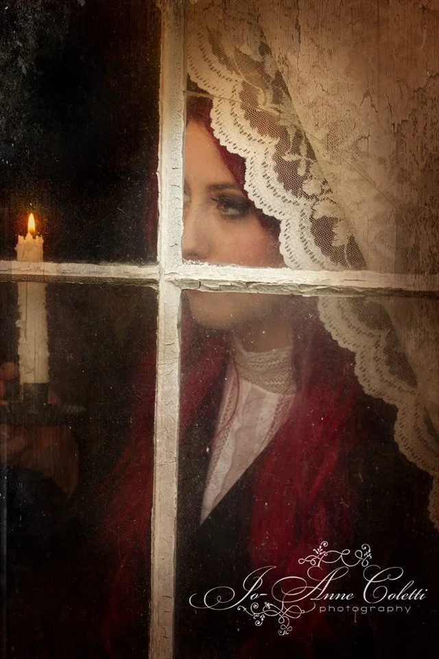 A woman with red hair seen through an old window with four panes, wearing a lace-trimmed veil and pearls, looking out thoughtfully as a candle flickers in the background.