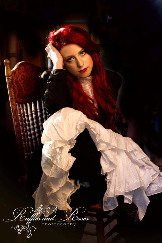 A woman with long red hair, wearing dark clothing, sitting on a wooden chair with a high back. She rests her head on her hand and looks directly at the camera. The lighting is warm and focused on her face, creating a dramatic effect. Text reads 'Ruff