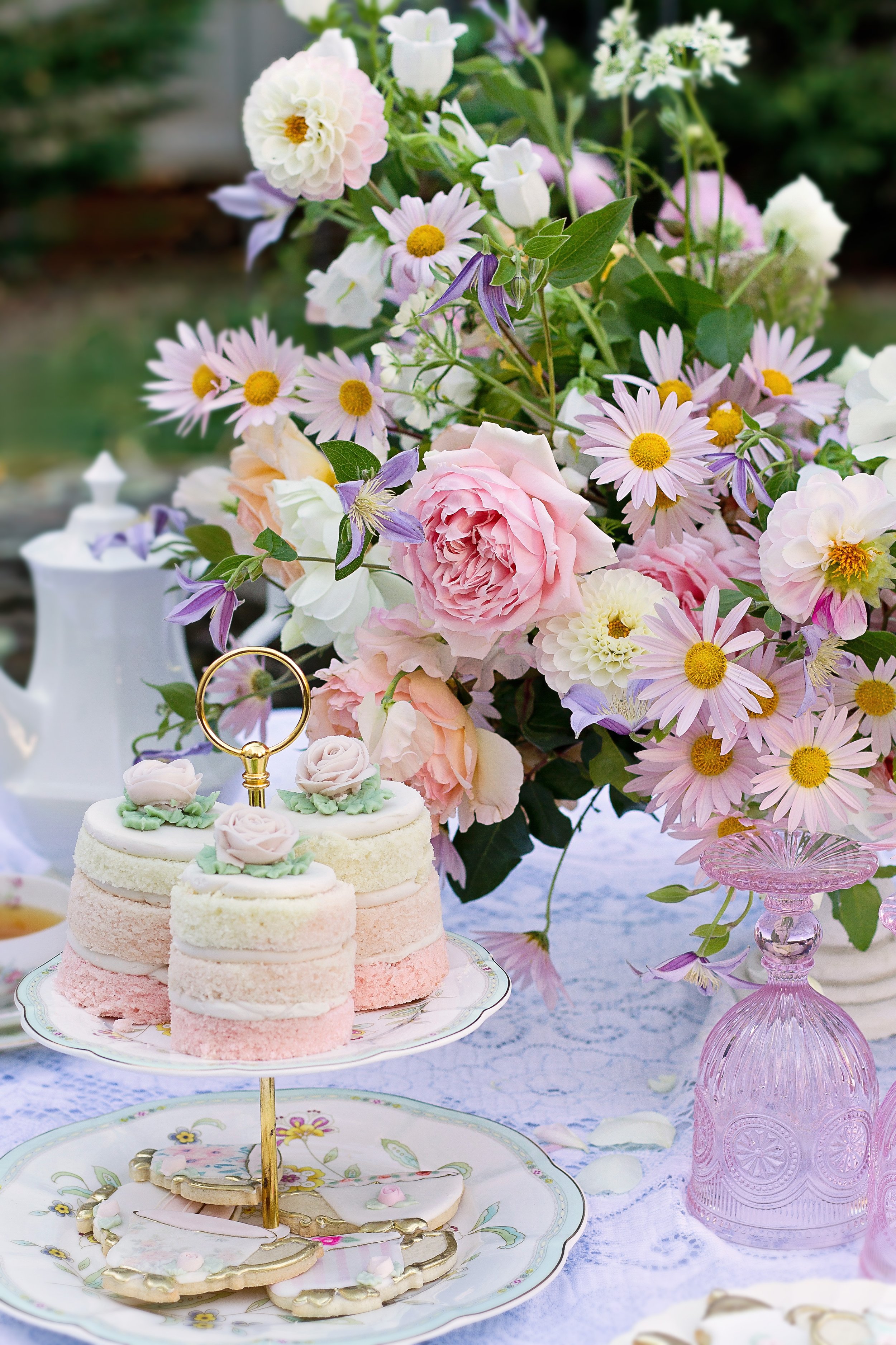 A floral arrangement with pink, white, and purple flowers on a table, with a three-tiered cake with pastel-colored layers and floral decorations, and dessert cookies with pink and white icing. The table is covered with a lace tablecloth and decorated