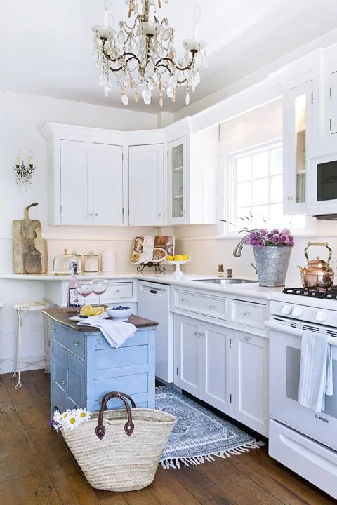 Bright white kitchen with cabinets, a small blue island, and flower arrangements on the counter, with natural light coming through a window.