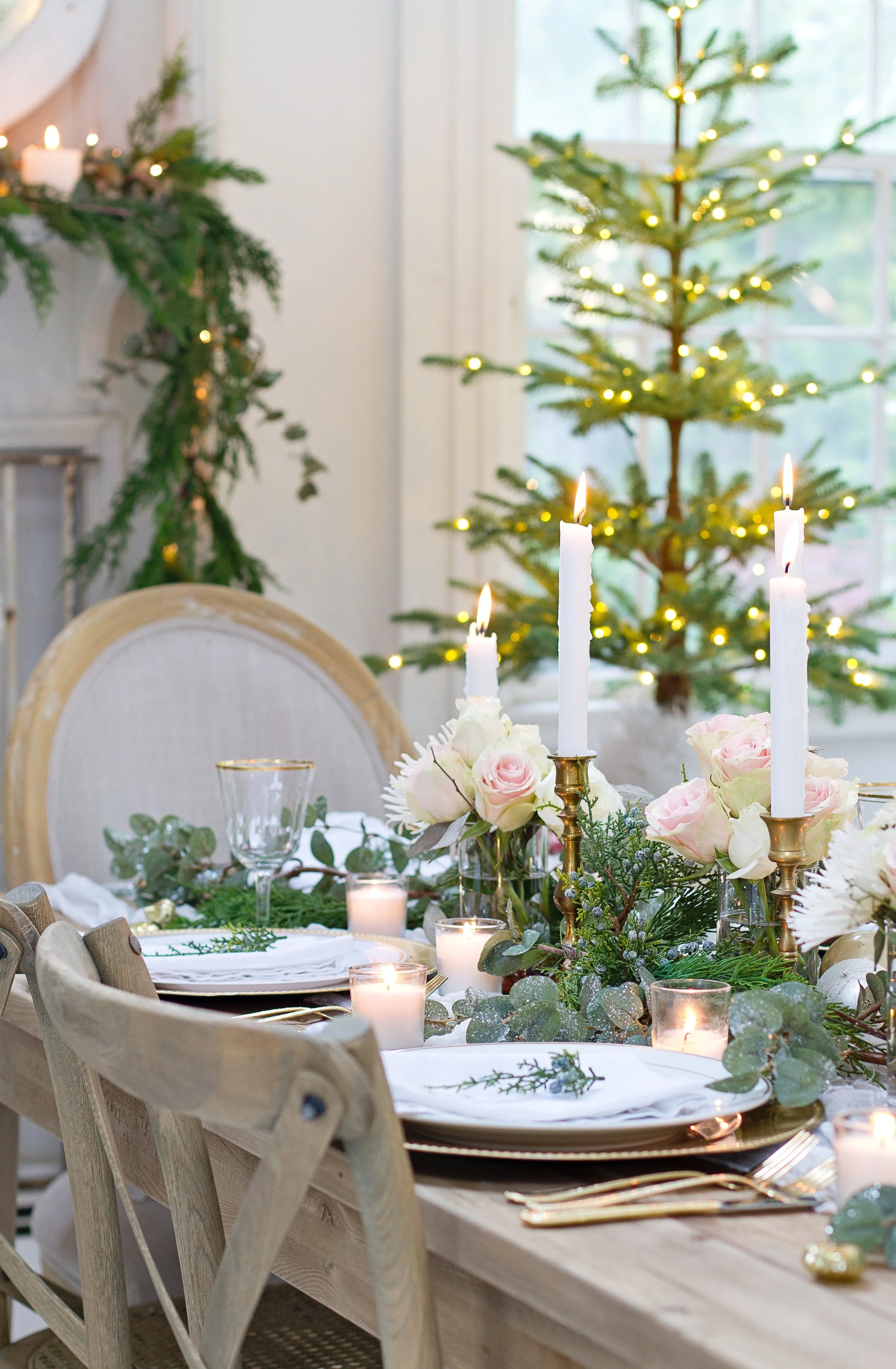 A Christmas decorated dining table with candles and floral arrangements, a Christmas tree with lights in the background, and a window letting in natural light.