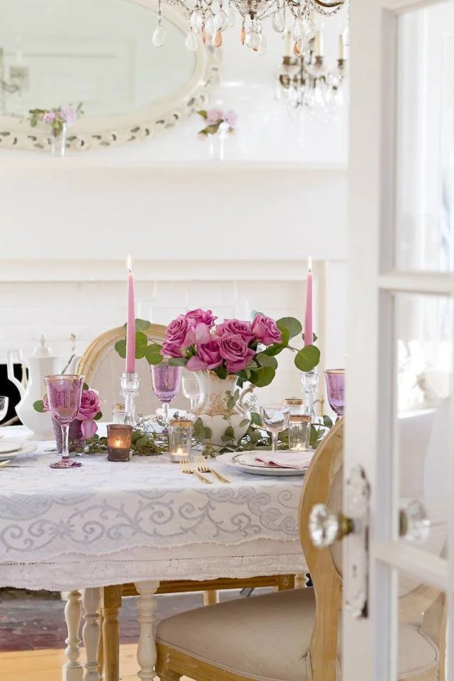 Elegant dining table set with pink candles, pink roses in the center, purple glasses, and gold flatware, in a bright room with a chandelier and a large mirror.