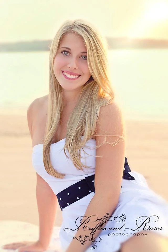 A young woman with long blonde hair and blue eyes smiling at the camera, sitting on a beach during sunset, wearing a white dress with a navy polka dot belt.