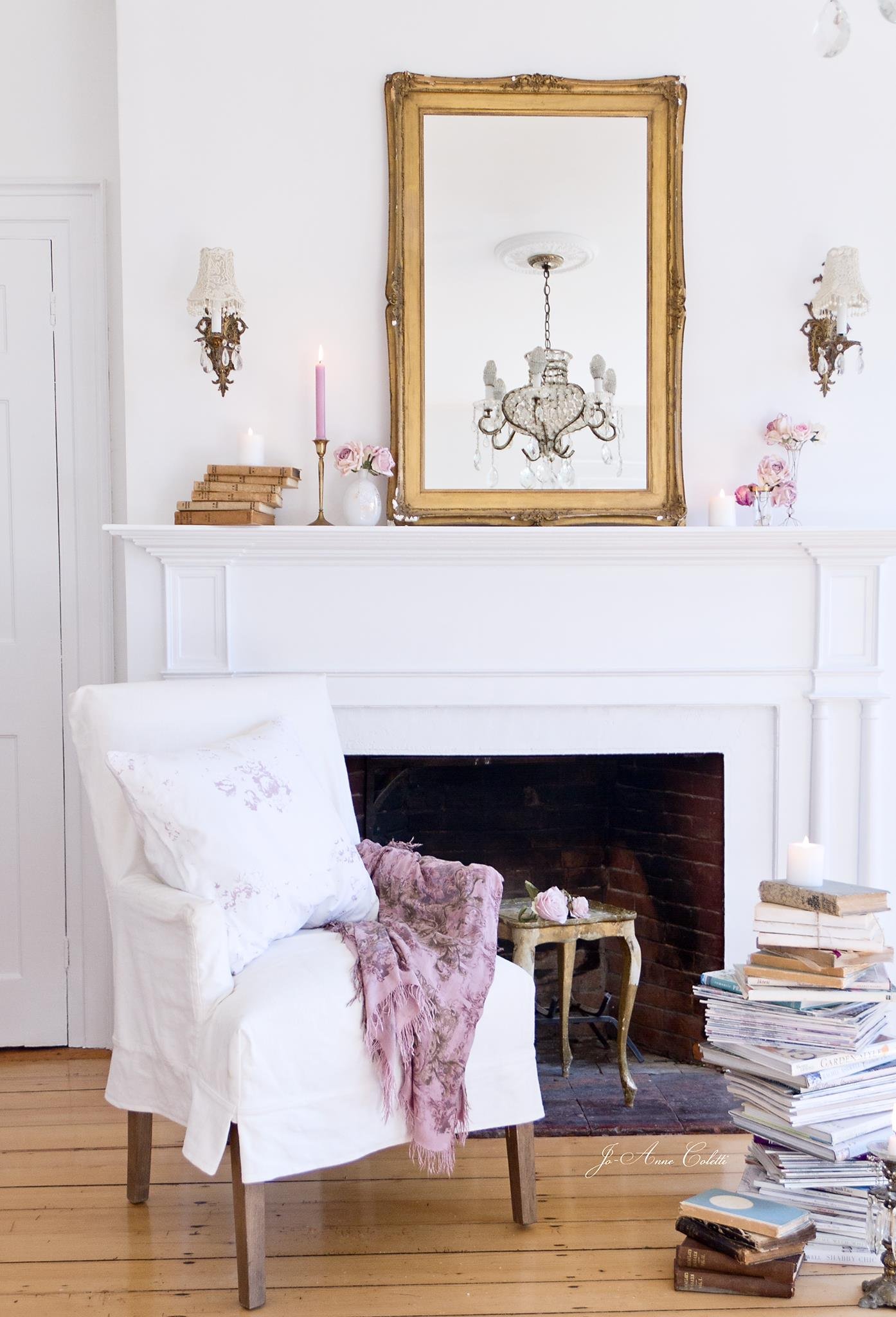 A cozy room with a white armchair draped with a pastel pink scarf, a side table filled with books, a large mirror above a white mantel, a vintage gold-Framed mirror, a crystal chandelier reflected in the mirror, wall sconces with lace shades, fresh p