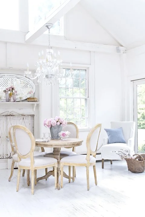Bright white living room with a round dining table, four chairs, a chandelier, a white armchair with a blue cushion, and large windows with greenery outside.
