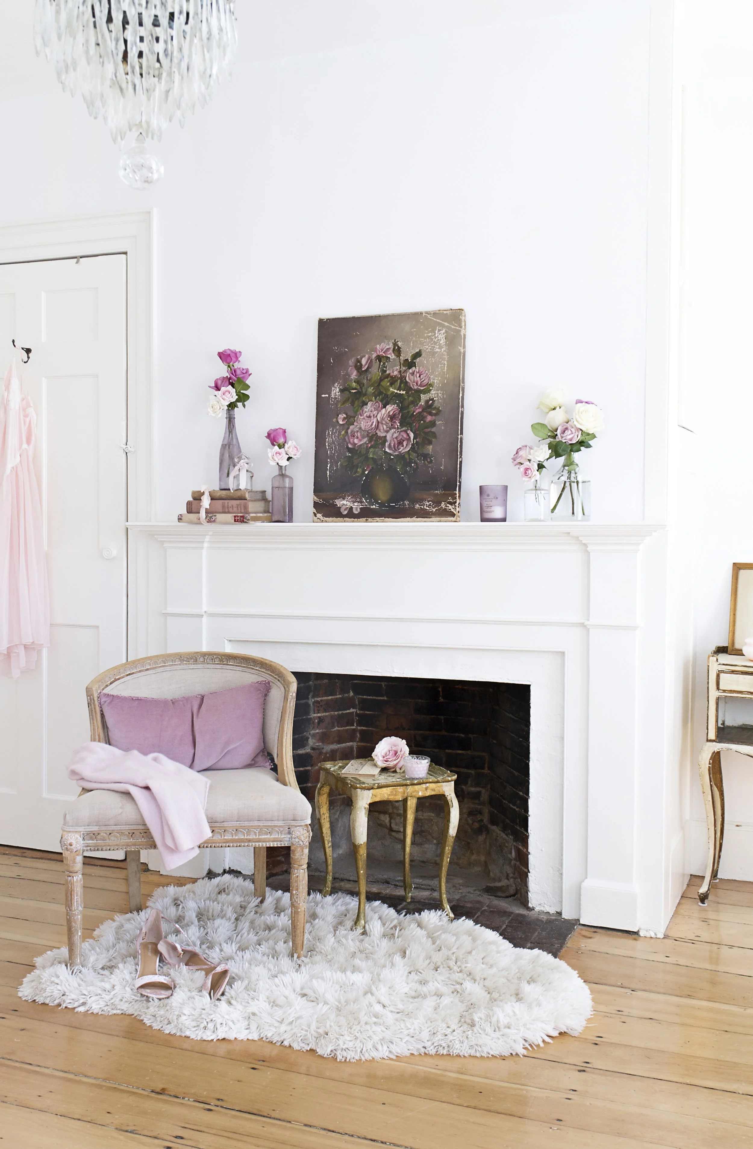 A cozy living room with a white painted brick fireplace, decorated with pink and white flowers in vases, books, and candles on the mantel. In front, there is a vintage-style armchair with pink velvet cushions and a pink blanket, a small gold side tab