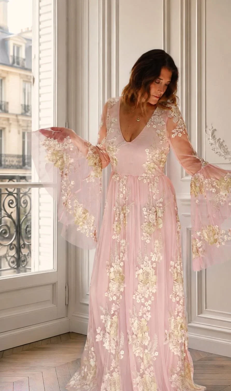 A woman in a sheer pink floral gown standing inside near a window, with French-style decor and a Parisian balcony visible outside.