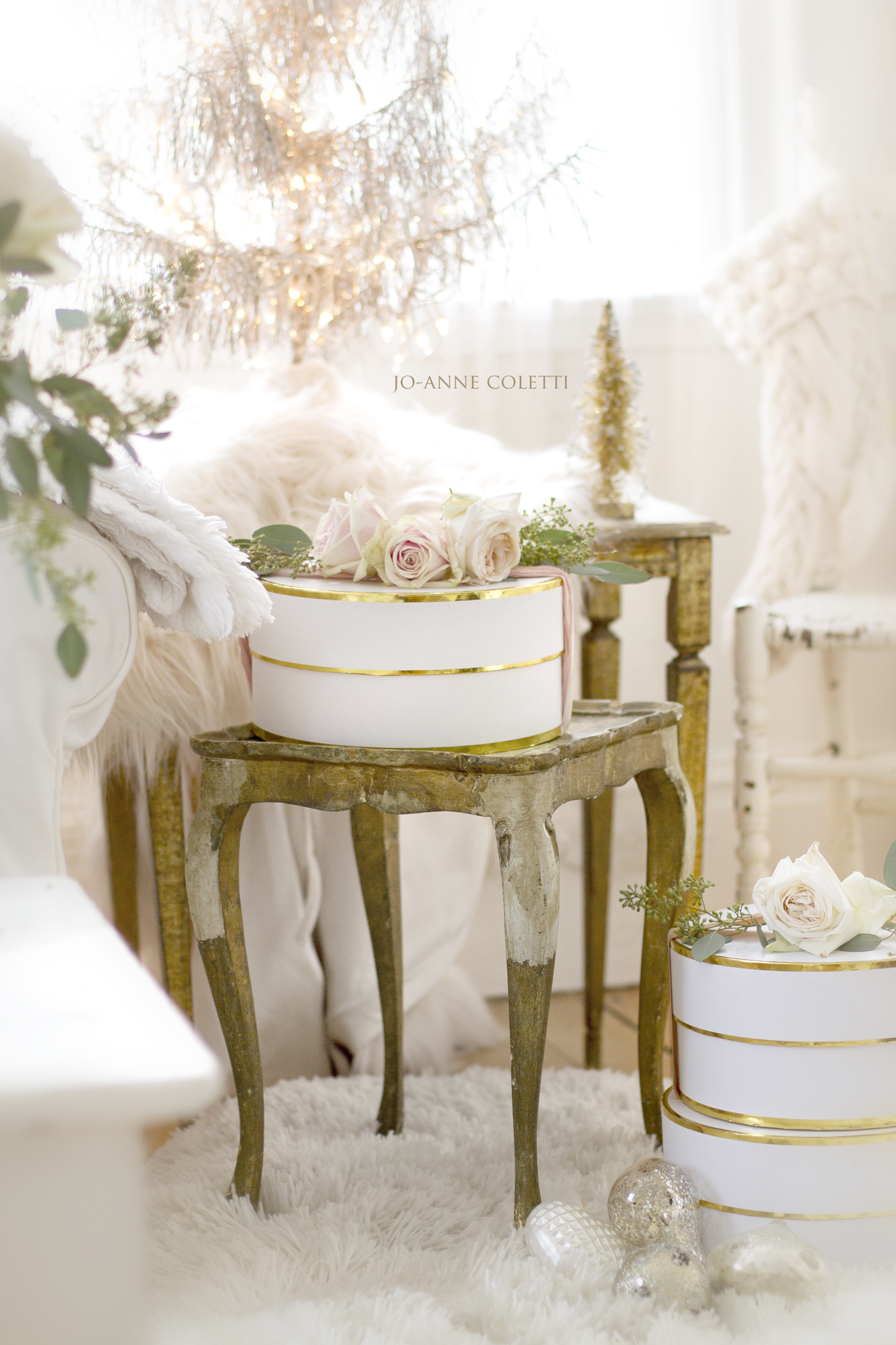 A cozy room decorated for winter with white and gold accents, including roses, a small Christmas tree, and decorative ornaments, all on rustic wooden tables and a fluffy white rug.