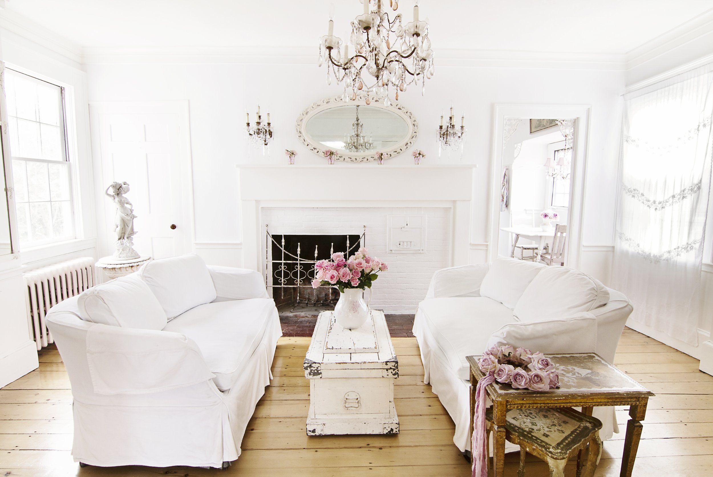 Living room with white sofas, a rustic coffee table with a bouquet of pink roses, white walls, a chandelier, an oval mirror above a fireplace, and a side table with a small bouquet of pink roses. Bright natural light fills the space.