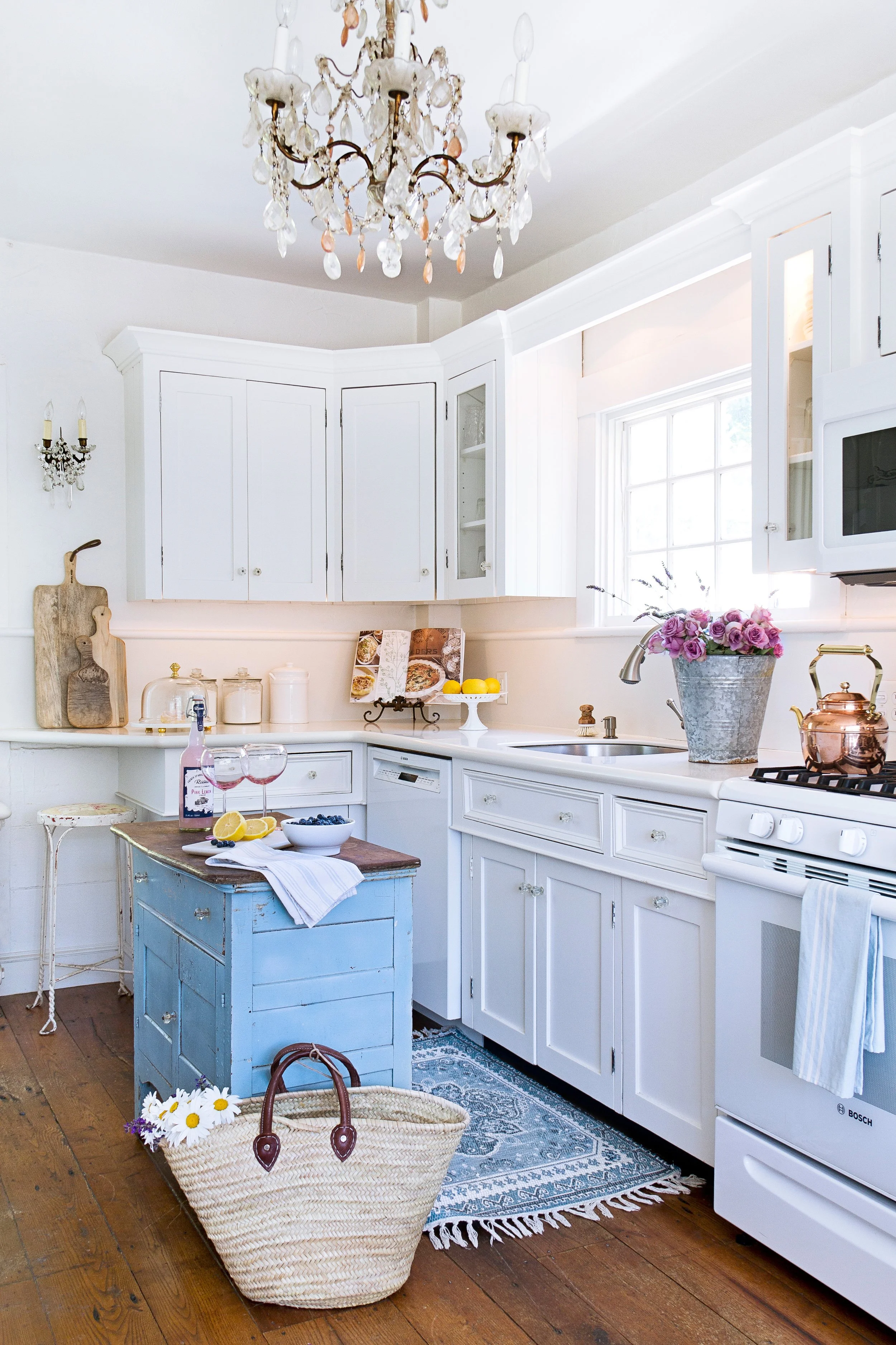 A bright kitchen with white cabinets, a chandelier, and a window. There is a small blue vintage cart with a white cloth, a basket with daisies, and a bottle of pink wine. The countertop has a vase with purple flowers, a copper teapot, and a cookbook.