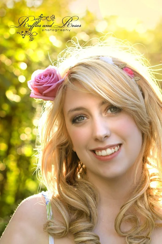 A young woman with blonde, curly hair and blue eyes smiling outdoors. She has a pink rose in her hair and a floral headband, with sunlight and greenery in the background.