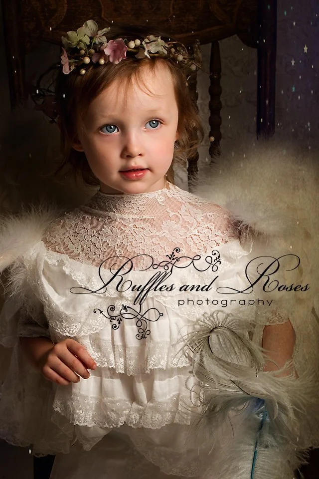 A young girl with curly hair and blue eyes wearing a white lace dress and a floral crown, holding a feather in her hand, in a softly lit room.